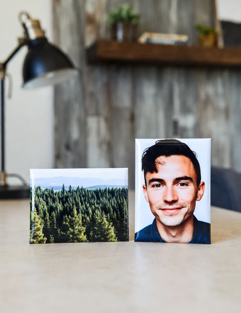 Two 2"x3" magnets on a desk, one with a forest and the other with a person, with a blurred indoor background.