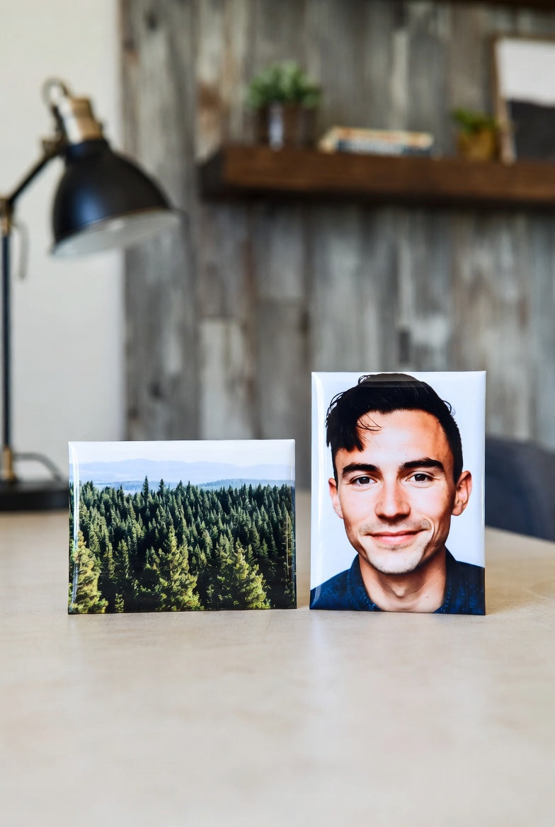Two 2"x3" magnets on a desk, one with a forest and the other with a person, with a blurred indoor background.