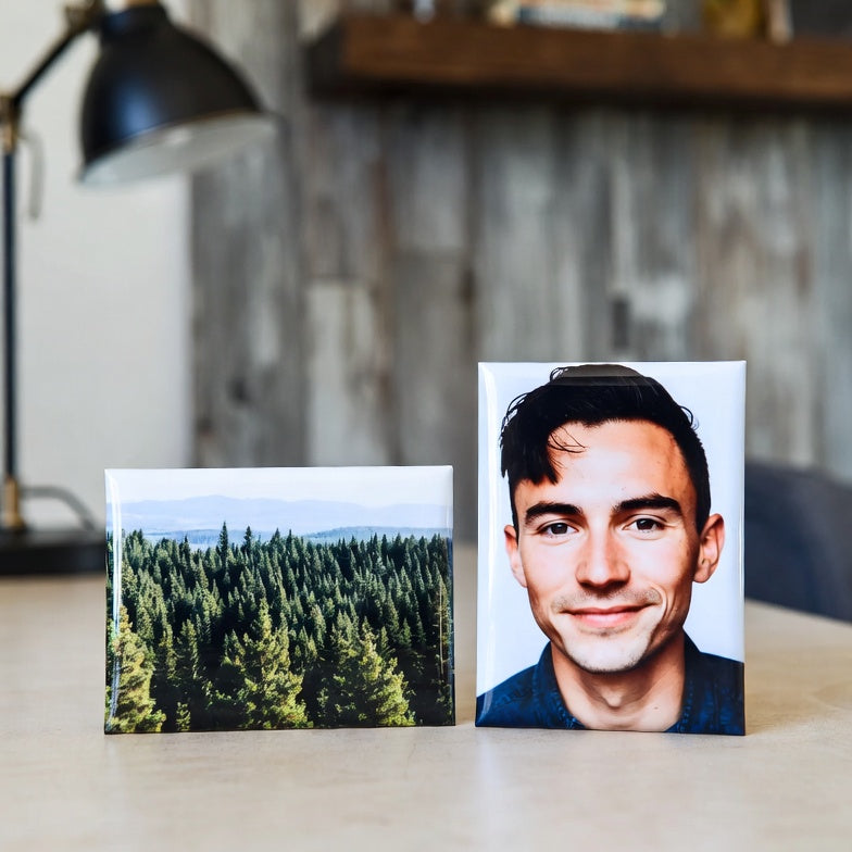 Two 2"x3" magnets on a desk, one with a forest and the other with a person, with a blurred indoor background.