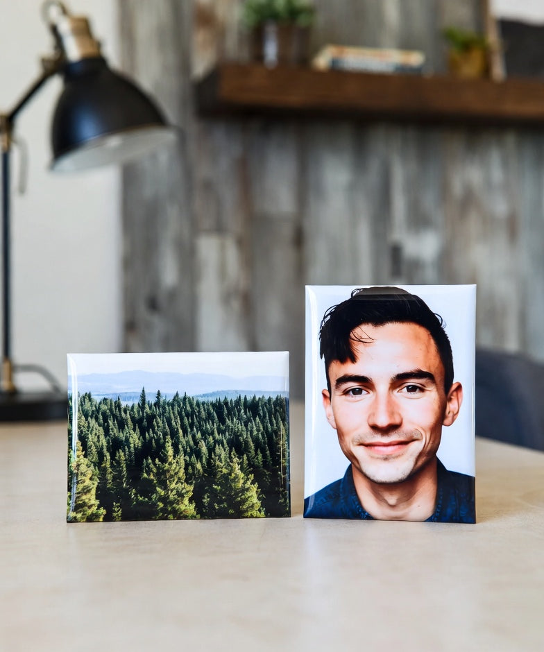 Two 2"x3" magnets on a desk, one with a forest and the other with a person, with a blurred indoor background.
